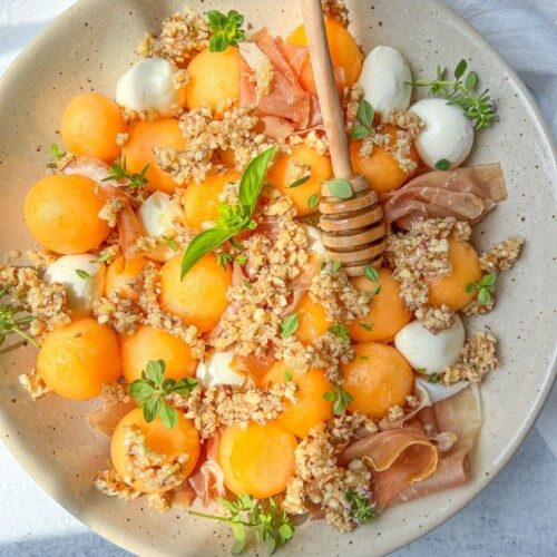 A speckled beige plate features prosciutto and melon balls, creamy cheese dollops, granola clusters, fresh herbs, and a honey dipper drizzled with honey for a savory-sweet delight.