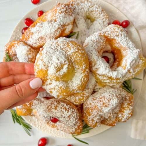 A hand holds a powdered sugar-covered zeppole above a plate piled with similar pastries, garnished with cranberries and rosemary, on a white surface.