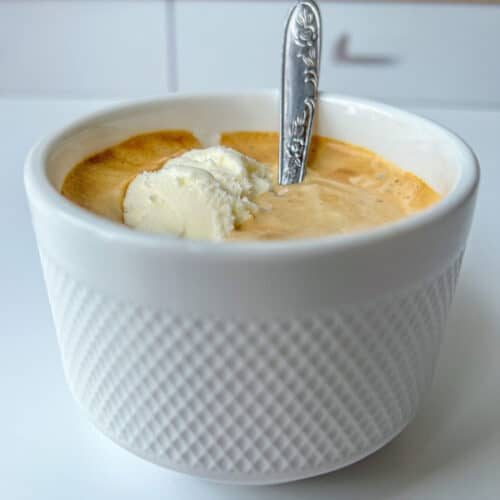 A white textured cup filled with coffee and a scoop of vanilla ice cream—a classic affogato recipe—finished with an ornate silver spoon. The cup sits on a white surface, the kitchen background softly blurred.