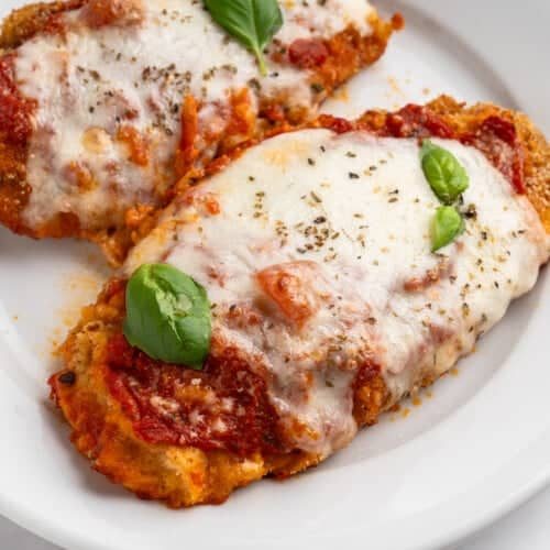 Two breaded chicken parmigiana covered in melted mozzarella cheese and tomato sauce, garnished with fresh basil leaves, served on a white plate.