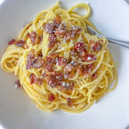 A plate of classic Carbonara topped with crispy pancetta and grated cheese, served on a white dish with a fork.