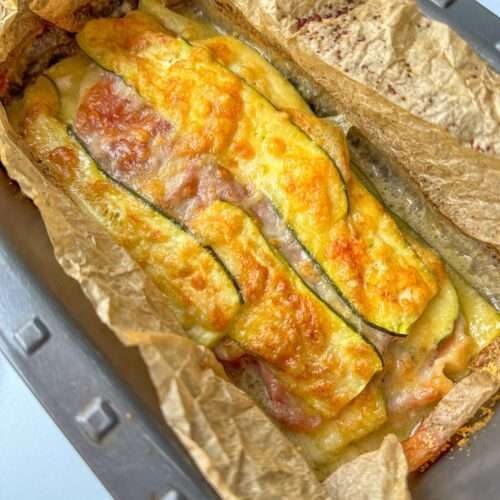 A Zucchini Lasagna Loaf with layers of zucchini and melted cheese is shown in a parchment-lined metal pan. The top is golden and slightly crispy, making this baked vegetable loaf irresistible.
