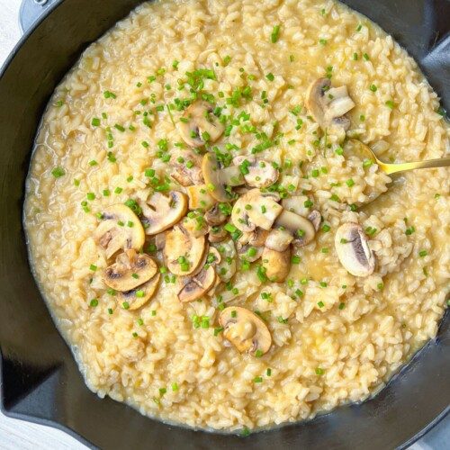 A black Staub pan filled with creamy mushroom risotto, made using a simple risotto recipe, is garnished with sautéed mushroom slices and chopped chives, and sits on a light wooden surface.