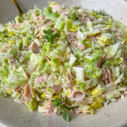 A close-up of a bowl filled with chopped cabbage, ham slices, and herbs, lightly seasoned with black pepper. A spoon rests in the bowl, showcasing this creamy and fresh Italian Chopped Salad.