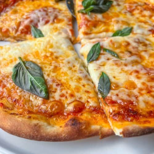 A close-up of an Easy Homemade Pizza topped with fresh basil leaves, cut into slices on a white plate with a pizza cutter nearby.