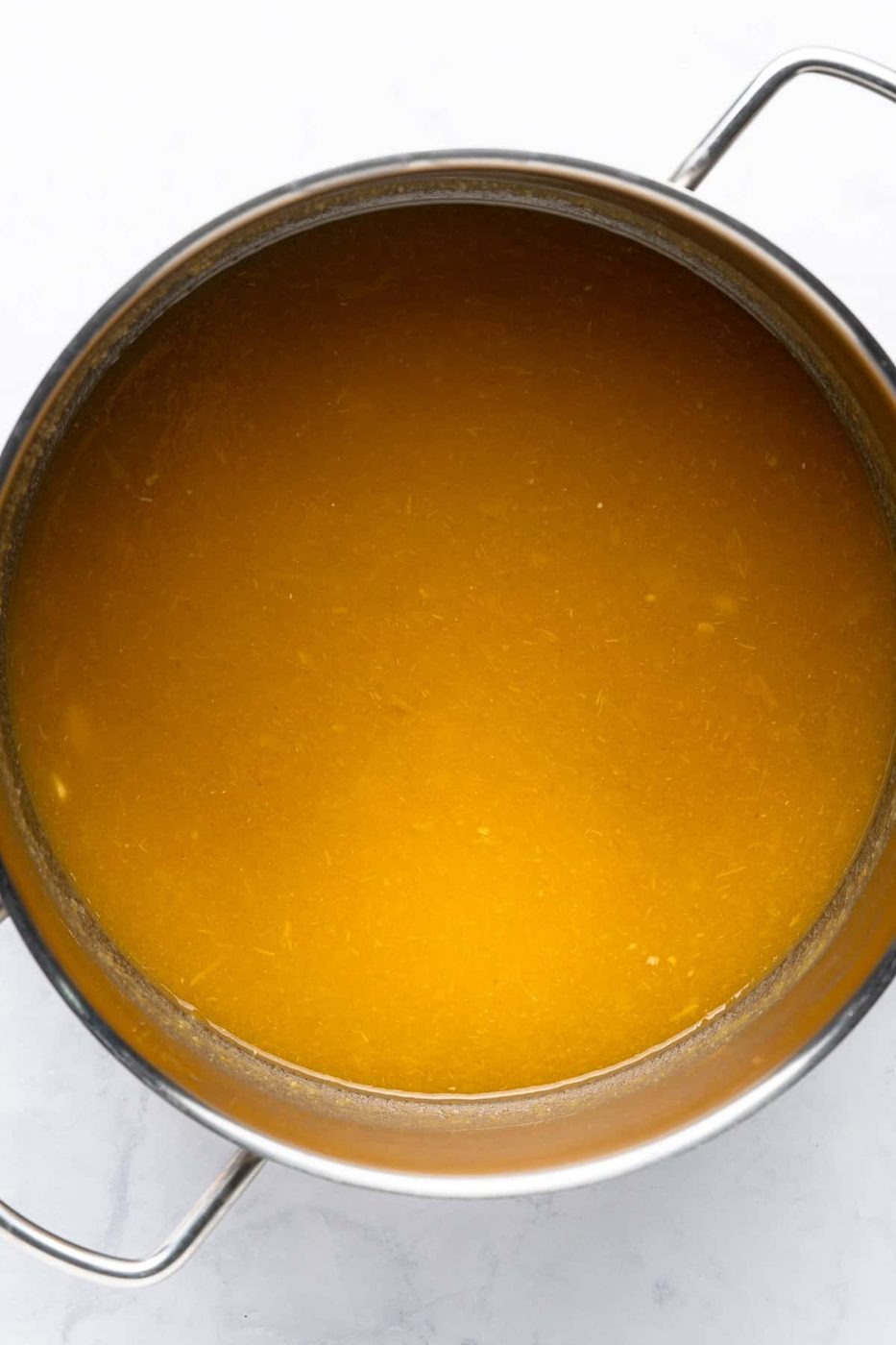 A large stainless steel pot filled with golden-brown Pastina Soup broth, placed on a light-colored surface. The broth appears smooth and rich, with a glossy sheen on the surface.