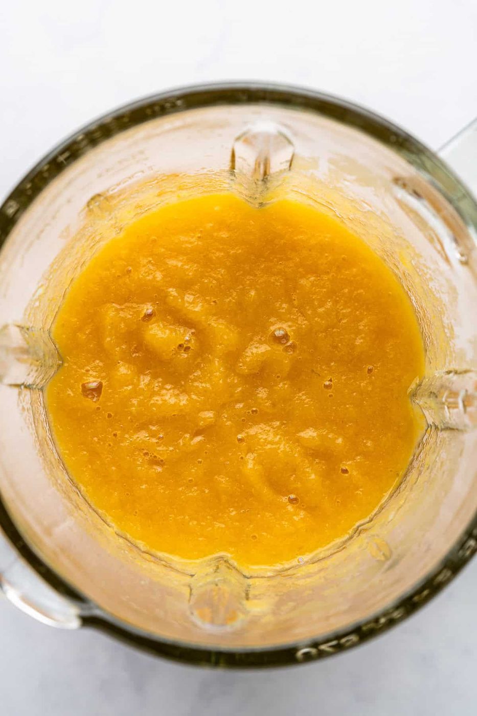 A top view of a blender containing smooth, bright orange mango puree. The glass sides of the blender show the creamy texture of the puree. The background is a light, subtly marbled surface.