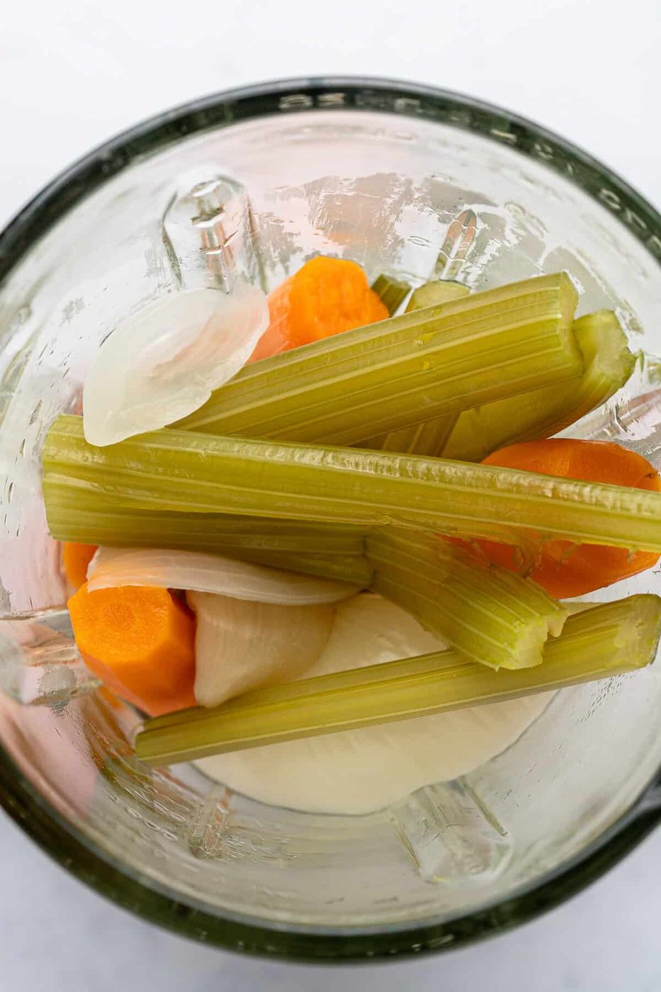 A glass blender filled with chopped celery stalks, sliced carrots, and onion pieces, viewed from above against a light background.