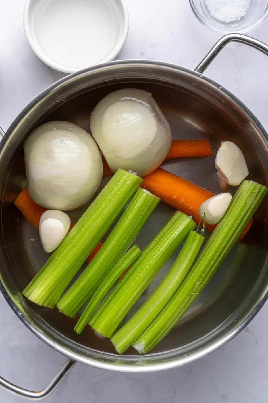A stainless steel pot containing water, halved onions, whole carrots, garlic cloves, and stalks of celery. The pot is on a light-colored surface, with a small bowl of salt and an empty bowl nearby.