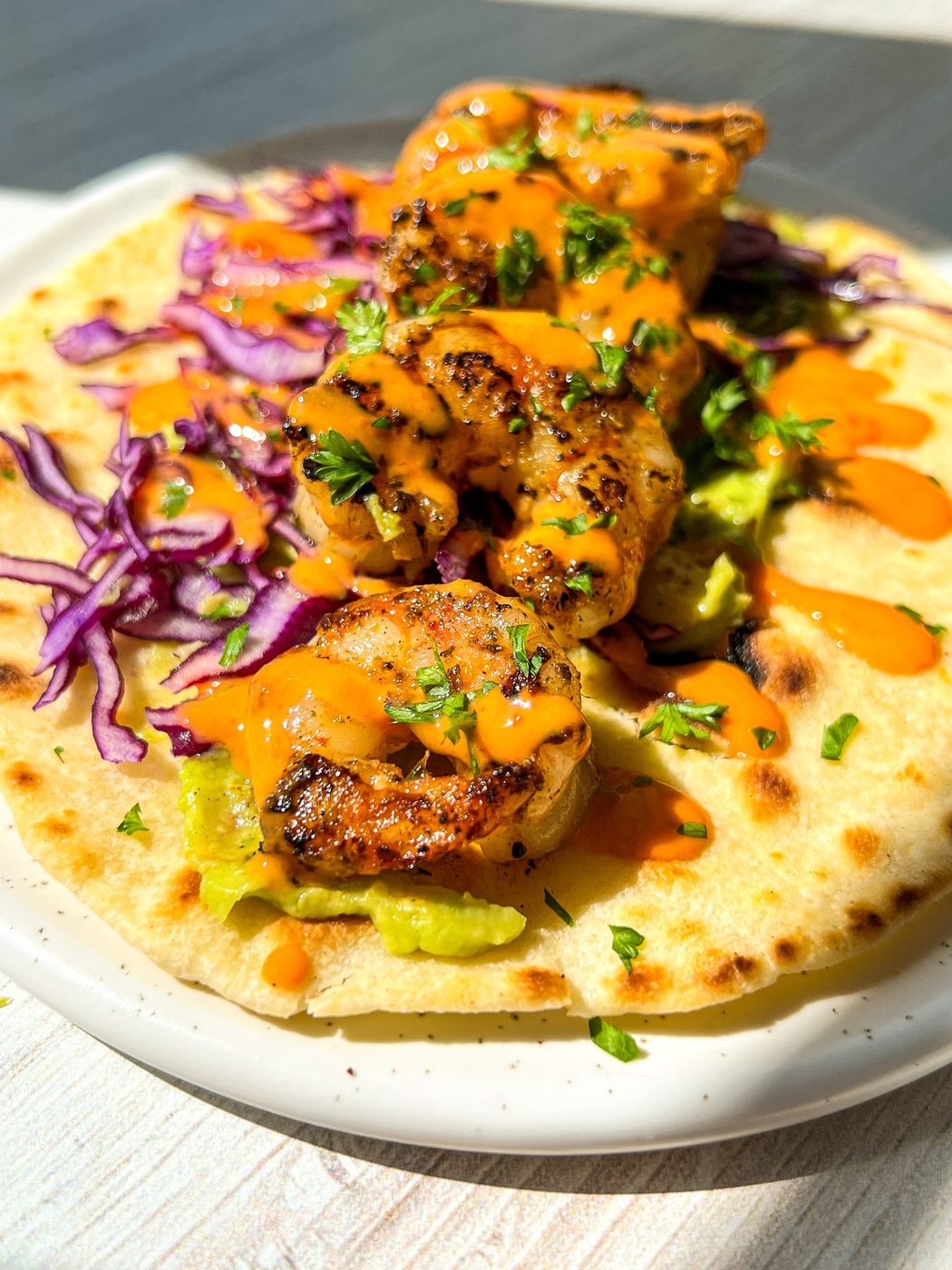 A grilled shrimp taco on a white plate, topped with vibrant red cabbage, avocado, and drizzled with orange sauce. The taco is garnished with chopped herbs, all set against a sunlit background.