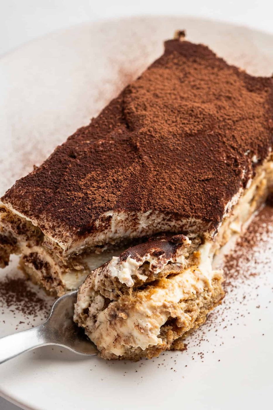 A slice of tiramisu on a white plate, dusted with cocoa powder. A spoon is scooping a portion from the dessert, revealing creamy layers.