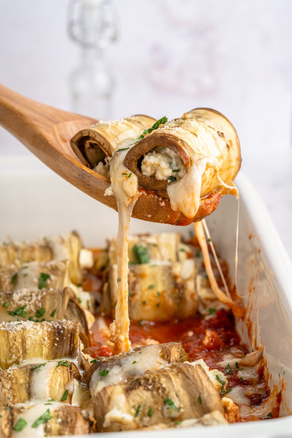 A wooden spoon lifts a serving of stuffed eggplant rollatini from a white baking dish. The rollatini is oozing melted cheese and garnished with chopped herbs. The dish is filled with more stuffed eggplant pieces in a rich tomato sauce, topped with melted cheese.