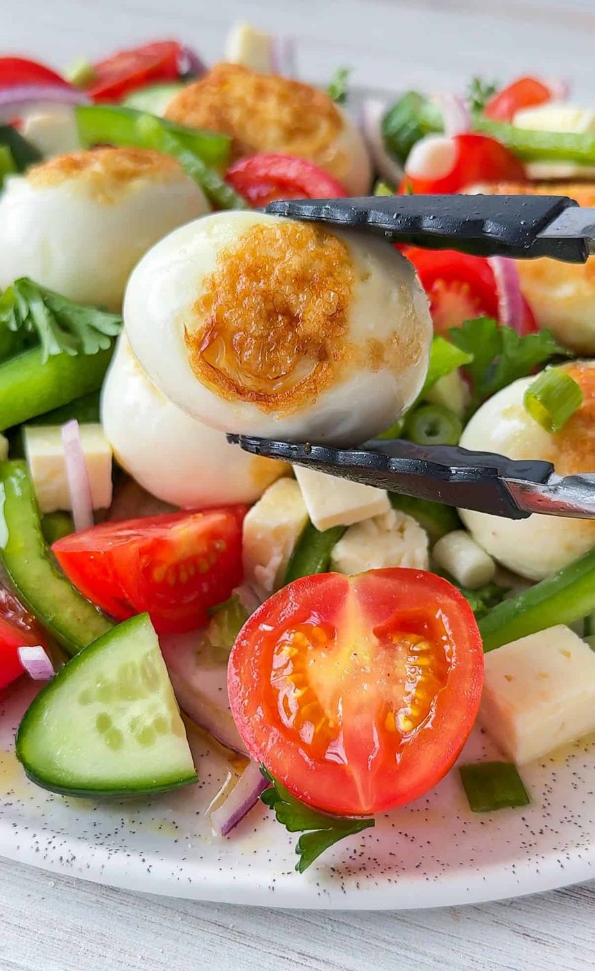 A close-up of a fresh salad featuring sliced cucumber, cherry tomatoes, red onion, green pepper, and cubes of cheese. Tongs hold a boiled quail egg with a golden-brown top, garnished with herbs.