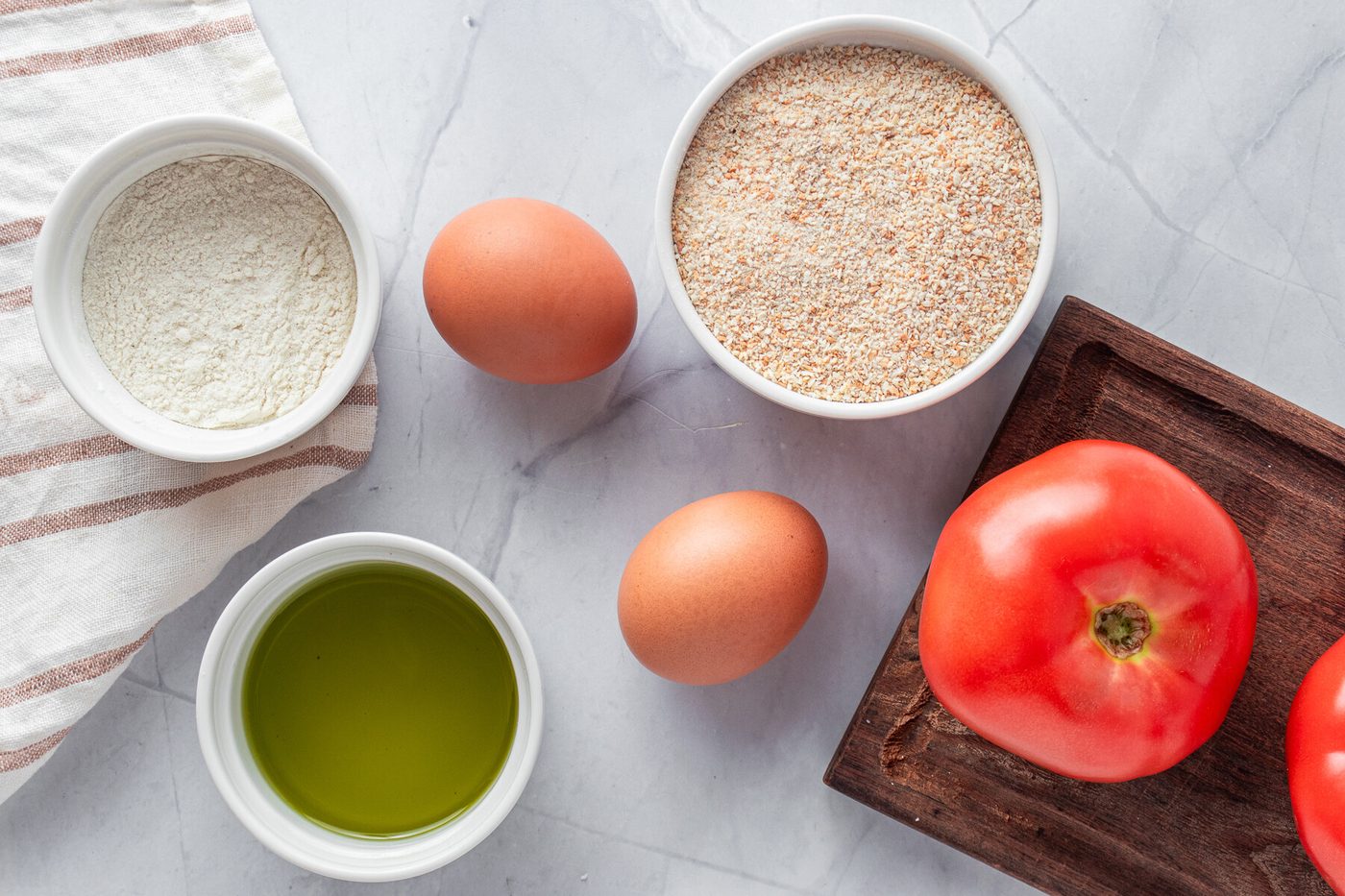 Ingredients for a recipe displayed on a marble surface: two large tomatoes on a wooden board, eggs, a bowl of breadcrumbs, olive oil in a small container, and flour in a white bowl with a striped cloth nearby.