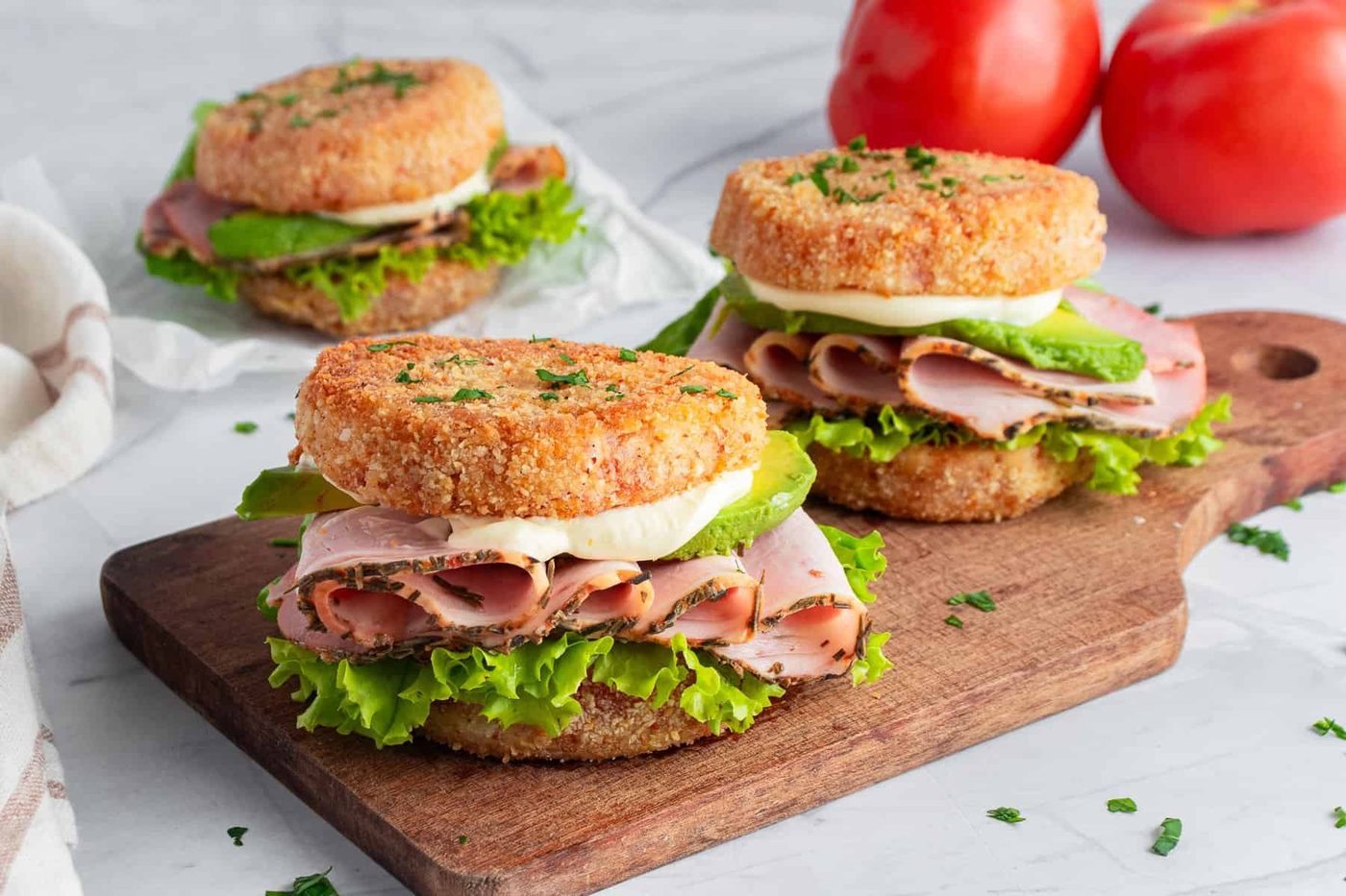Three sandwiches with deli meat, lettuce, avocado, and mayonnaise are stacked between two crispy breaded rounds on a wooden board. 