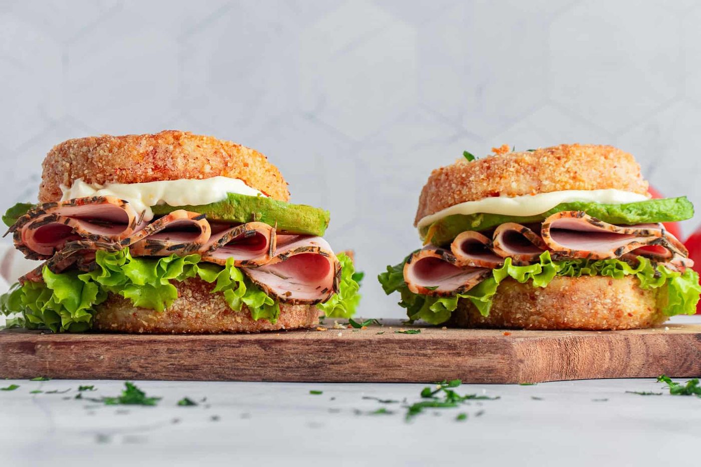 Two sandwiches made with crispy breaded buns, filled with layers of lettuce, sliced meat, avocado, and topped with white sauce, are placed on a wooden board. The background features a subtle hexagonal pattern.