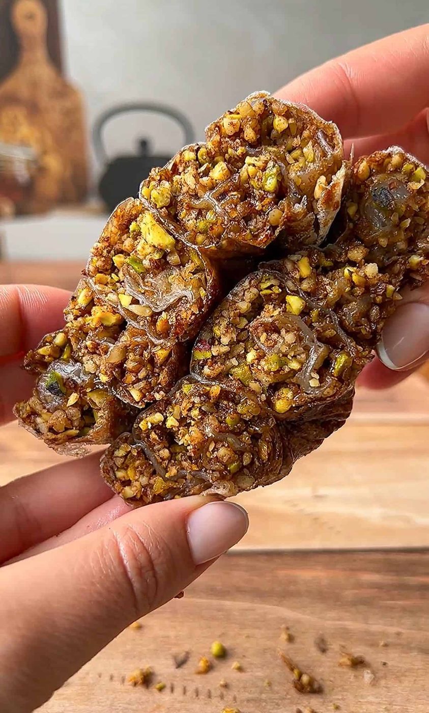 Close-up of hands holding a cluster of date rolls stuffed with chopped pistachios and nuts. The texture is rough and nutty, and the rolls are segmented, revealing the dense filling inside.