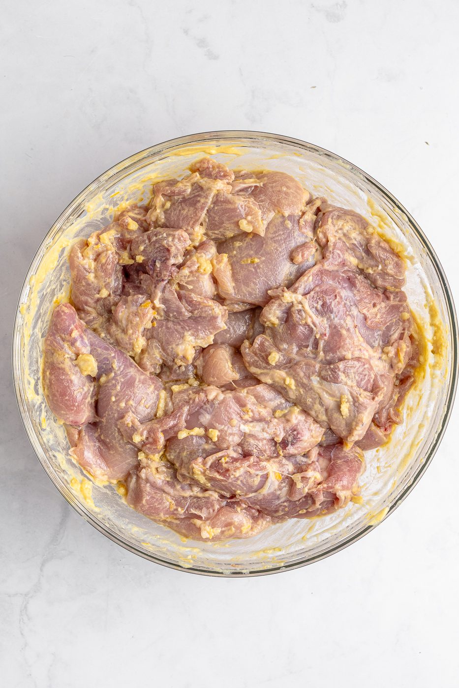 A bowl of raw marinated chicken pieces in a light yellow marinade, placed on a white marble surface. The marinade appears to contain visible chunks of seasoning or spices.