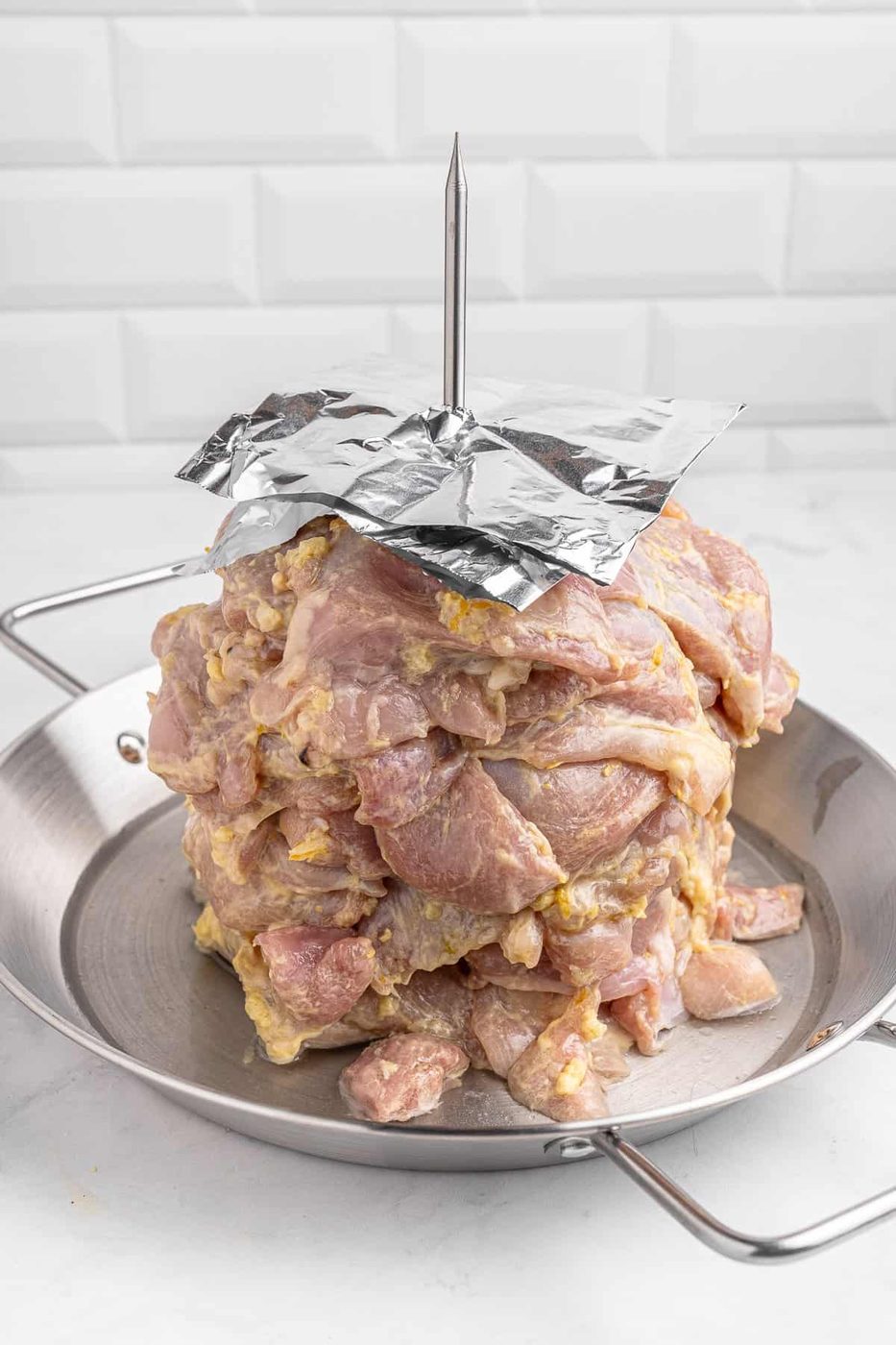 Raw marinated meat stacked vertically on a skewer in a metal tray, with a foil sheet covering the top. The background features white tiled walls.