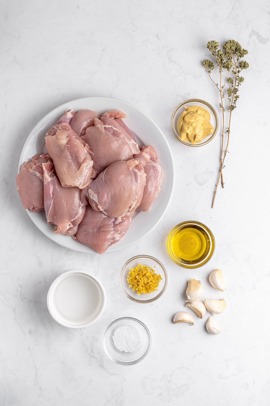 Raw chicken thighs on a white plate, surrounded by small bowls of mustard, olive oil, milk, lemon zest, and salt. Several garlic cloves and sprigs of dried herbs are placed on a white marble surface.
