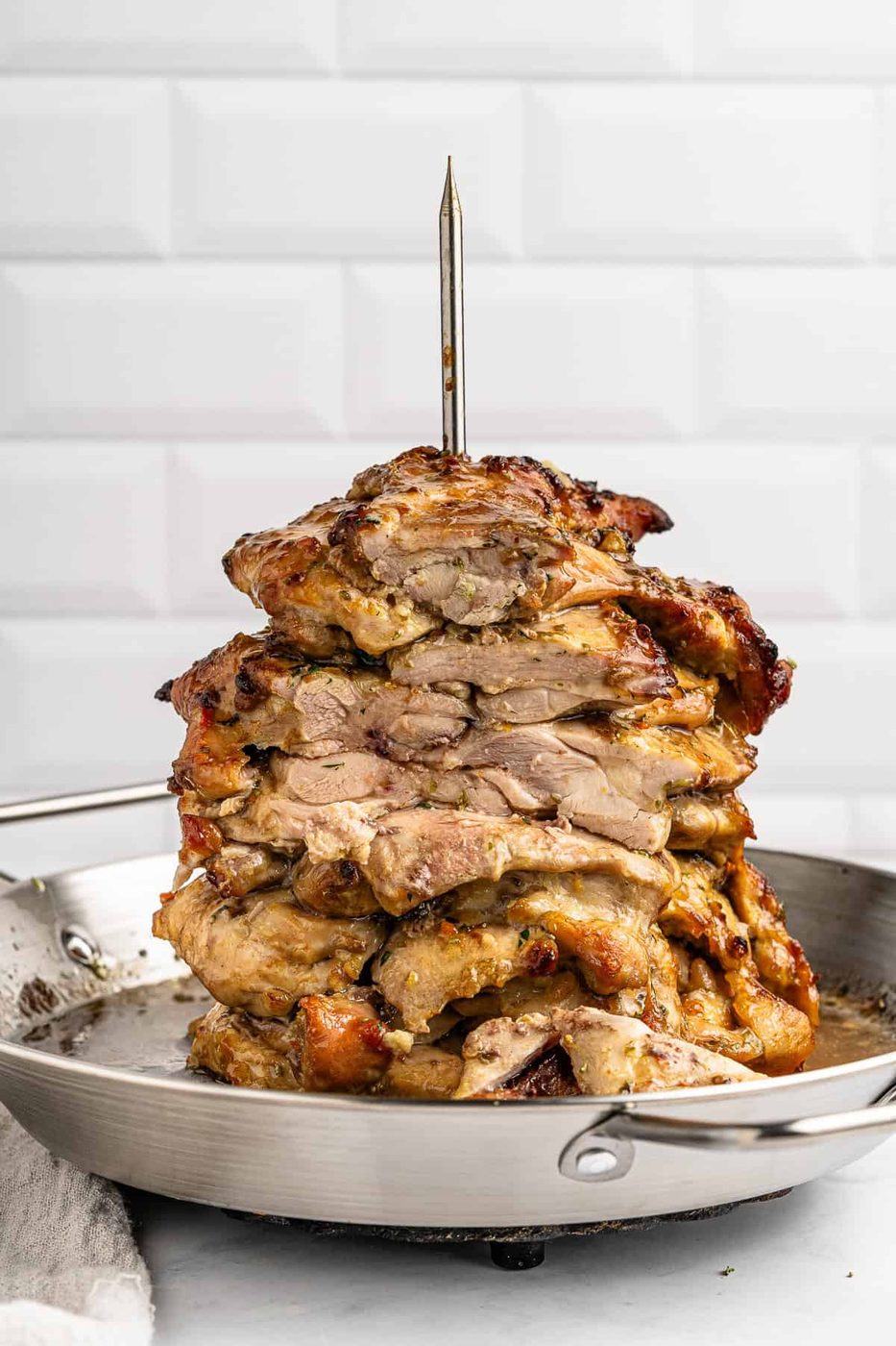Stacked layers of marinated, grilled meat on a vertical skewer in a metal pan, set against a white tiled background.