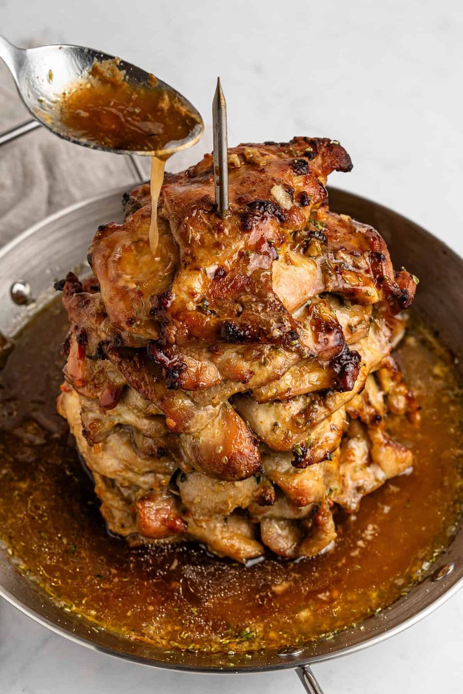 A vertical stack of marinated, grilled meat on a skewer in a pan, with a spoon drizzling sauce over it. The meat is browned and juicy, sitting in a pool of sauce. The background is a light, neutral countertop.
