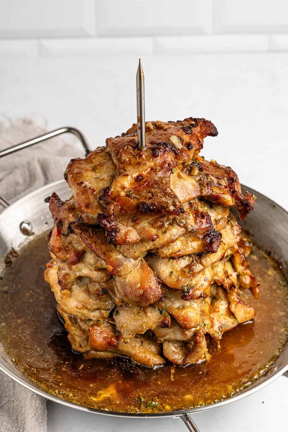 A stack of marinated and grilled meat pieces is skewered vertically on a spit in a round metal pan. The meat is browned and juicy, surrounded by flavorful juices. A beige cloth napkin is partially visible on the left side.