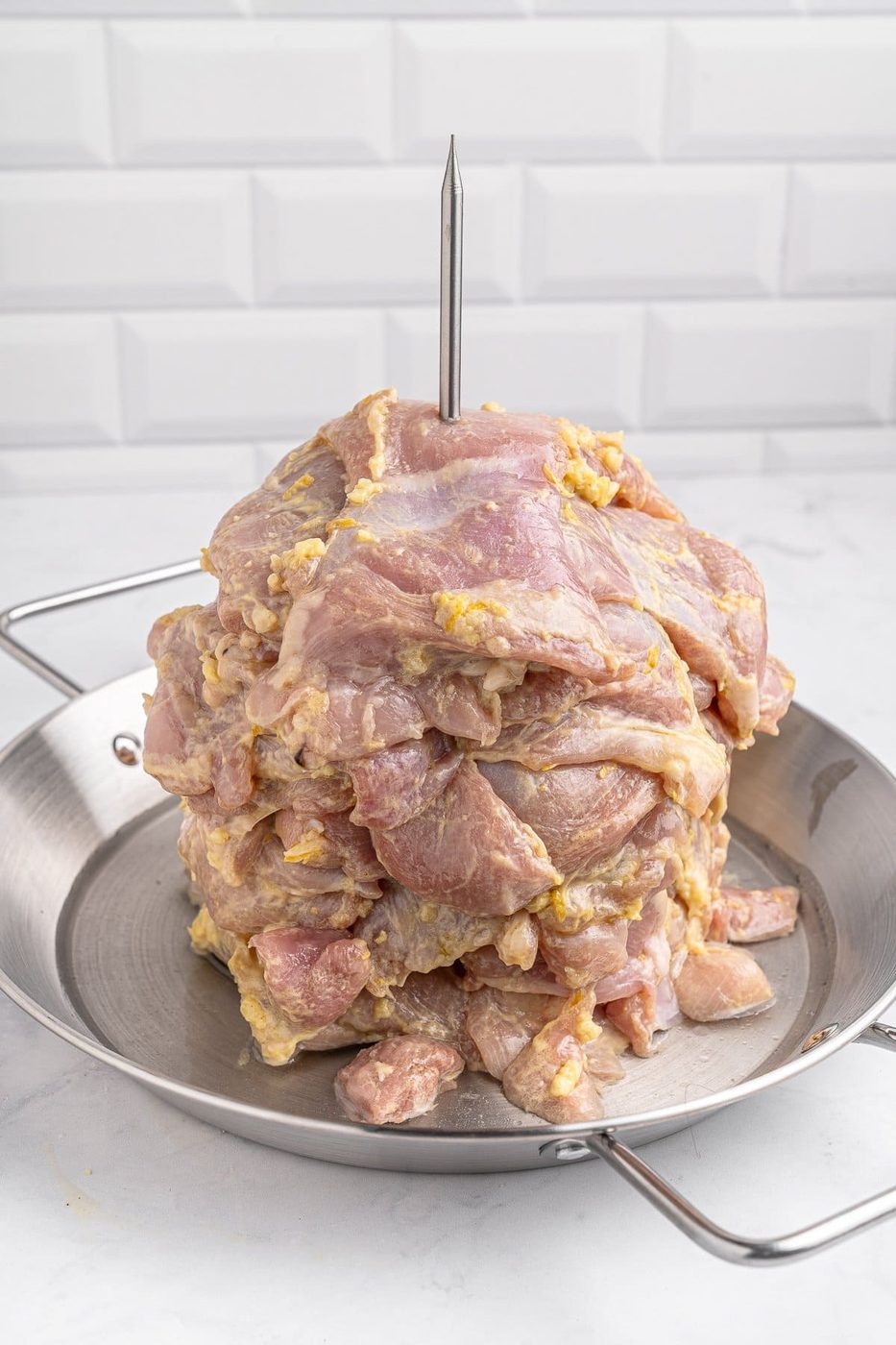 Raw marinated meat stacked on a skewer, placed vertically on a metal pan, ready for grilling. The background features white tiled walls.