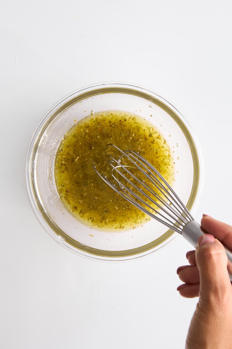 A hand holding a whisk mixing a light green, herb-speckled liquid in a clear glass bowl on a white surface.