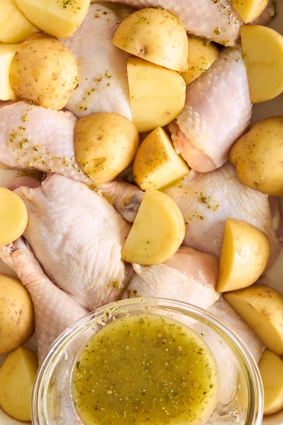 Raw chicken legs and halved yellow potatoes arranged in a baking dish, with a bowl of seasoned olive oil marinade placed on top, ready to be poured over the ingredients.