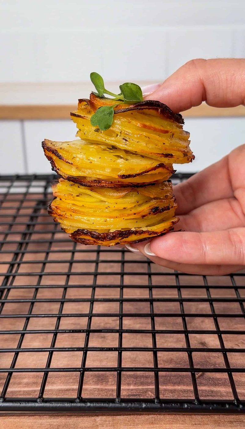 A hand holds a stack of crispy, golden greek lemon potato stacks, thinly sliced and garnished with a sprig of green herb, above a black cooling rack on a wooden surface.