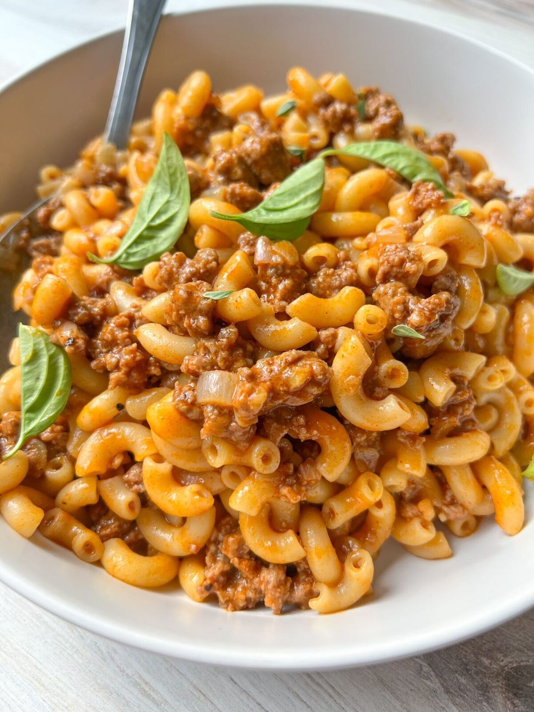 A white bowl filled with creamy macaroni beef pasta garnished with fresh basil leaves. The pasta is coated in a rich, savory sauce and chunks of ground beef are mixed throughout. A silver fork rests on the side of the bowl.