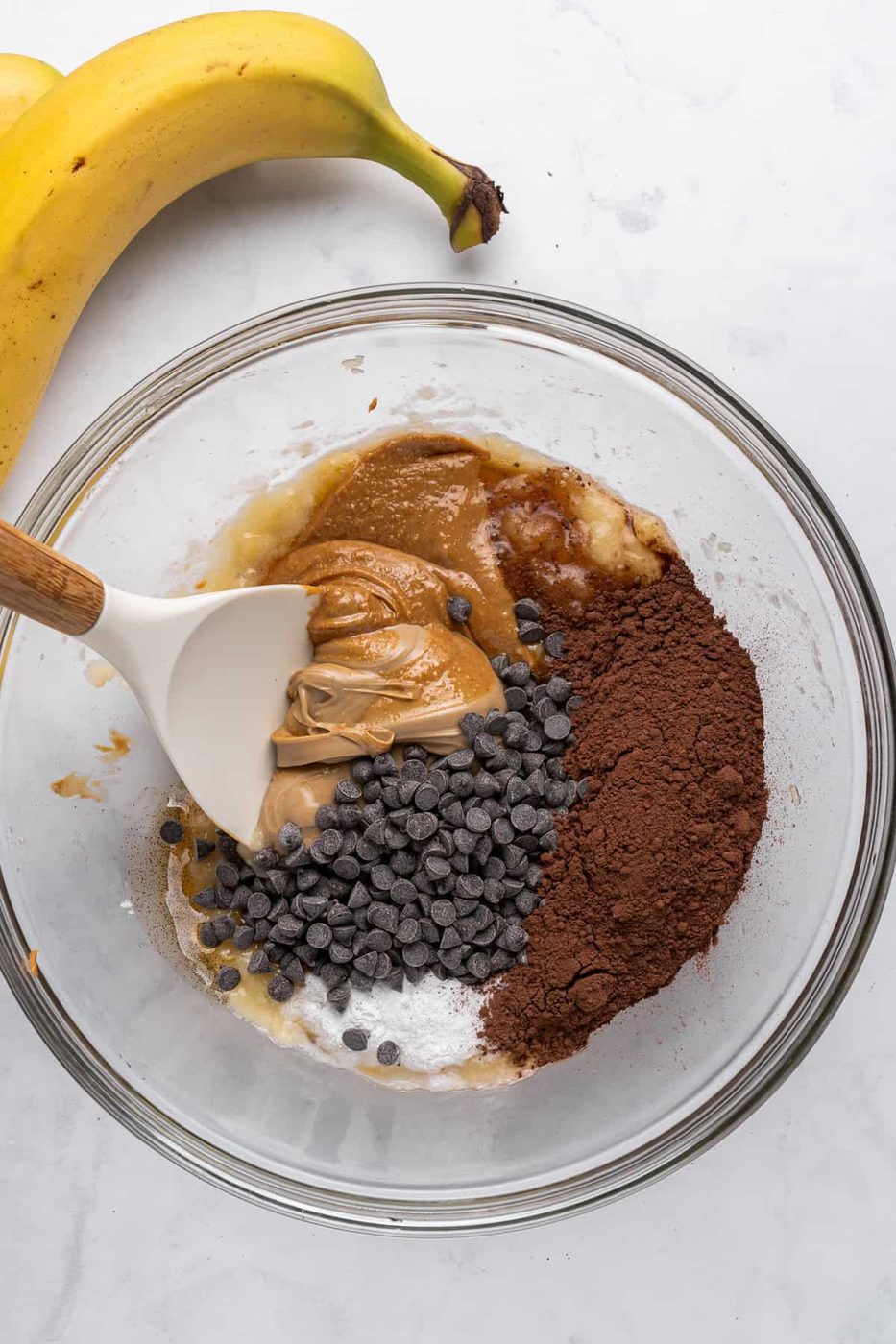 A glass bowl contains ingredients for baking: mashed banana, peanut butter, chocolate chips, cocoa powder, and a white spatula mixing them. A whole banana is placed beside the bowl on a marble surface.