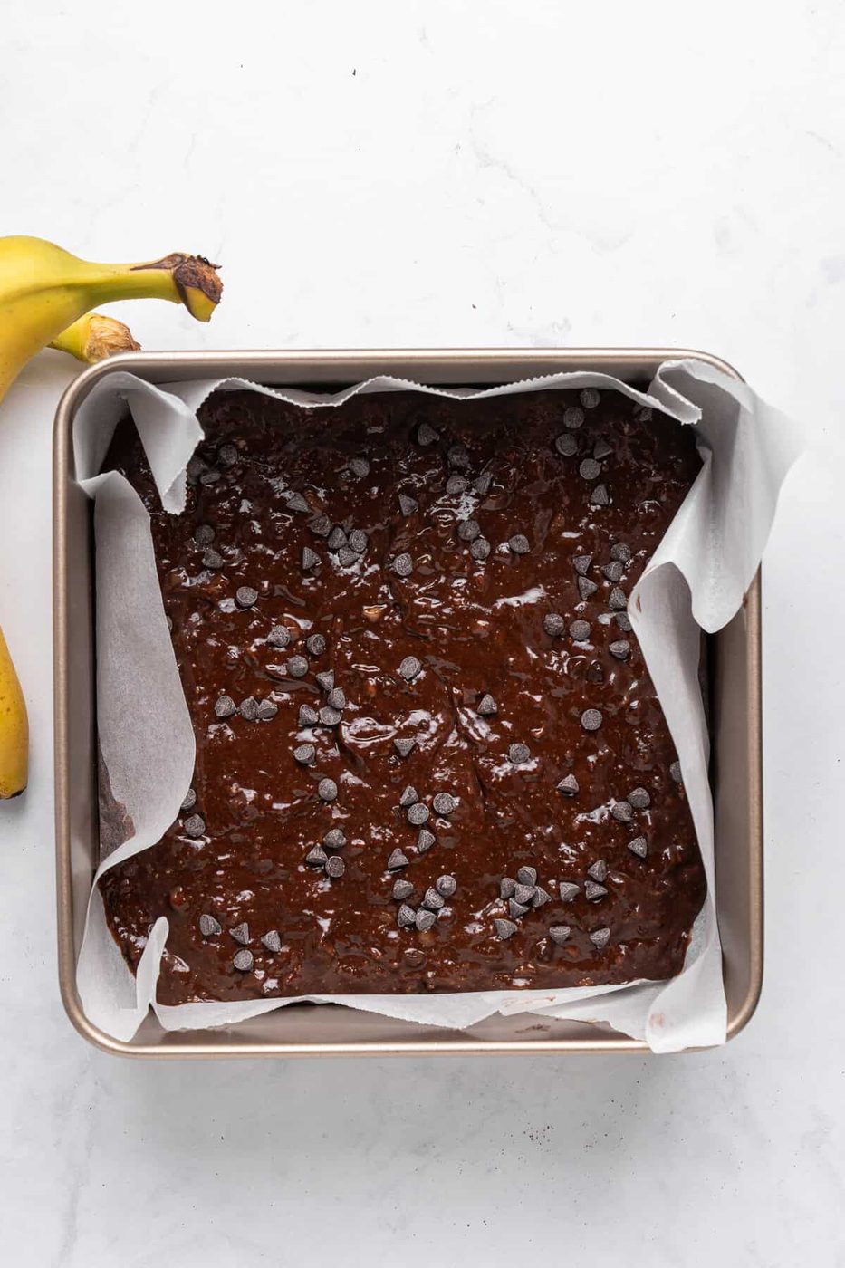 A square baking pan filled with raw chocolate brownie batter topped with chocolate chips, lined with parchment paper. Two ripe bananas are partially visible on the side on a white marble surface.