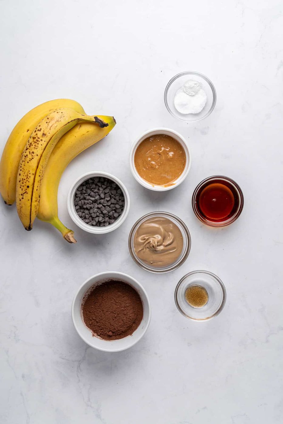 Ingredients for a baking recipe arranged on a marble surface: two bananas, small bowls of chocolate chips, cocoa powder, almond butter, peanut butter, a glass of maple syrup, and smaller bowls with baking soda and vanilla extract.
