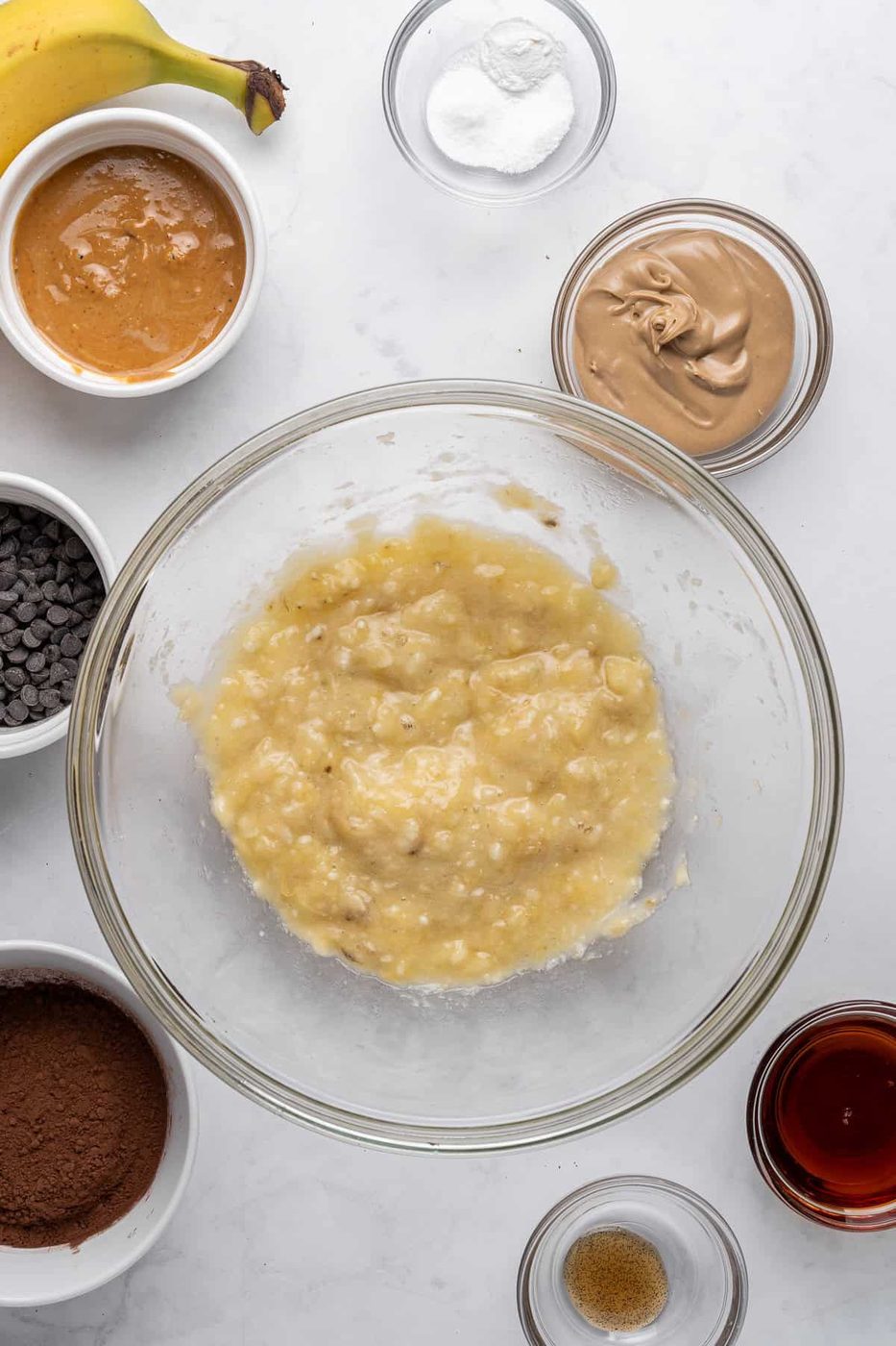 A glass bowl with mashed bananas on a marble surface, surrounded by ingredients in small bowls: peanut butter, chocolate chips, cocoa powder, a banana, baking soda, salt, vanilla extract, and brown sugar.