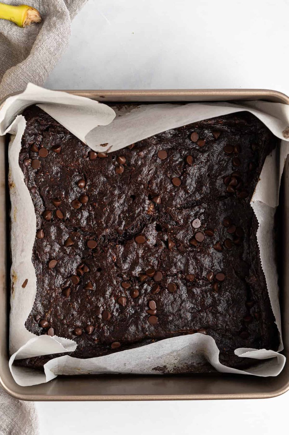 A square baking pan lined with parchment paper containing freshly baked chocolate brownies. The brownies are dark and topped with chocolate chips. The surface appears rich and moist. A beige fabric is partially visible on the side.