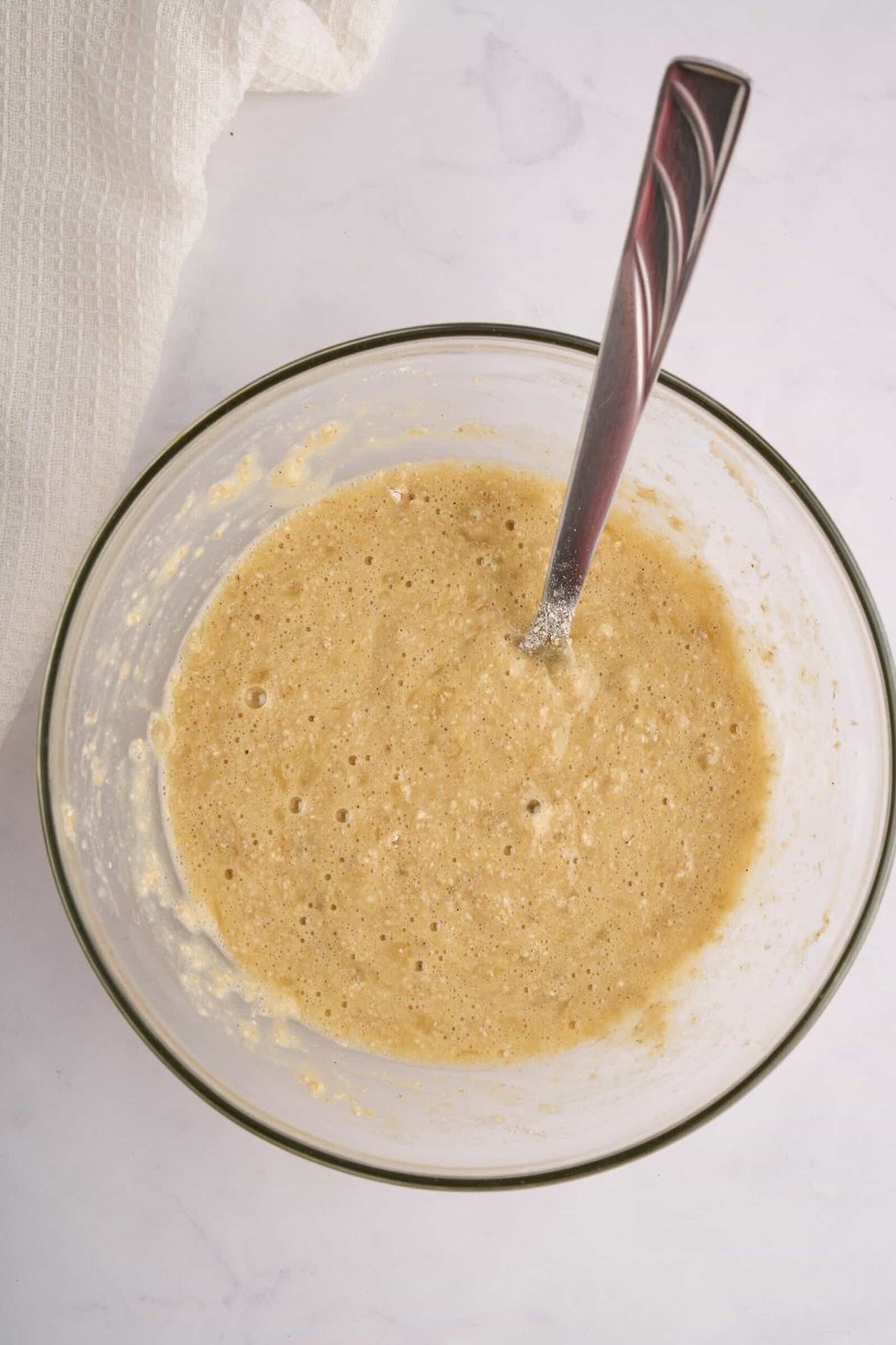 A glass bowl filled with a light brown cake batter and a silver spoon resting in it. The bowl is placed on a white marble countertop, and a white cloth is partially visible in the top left corner.