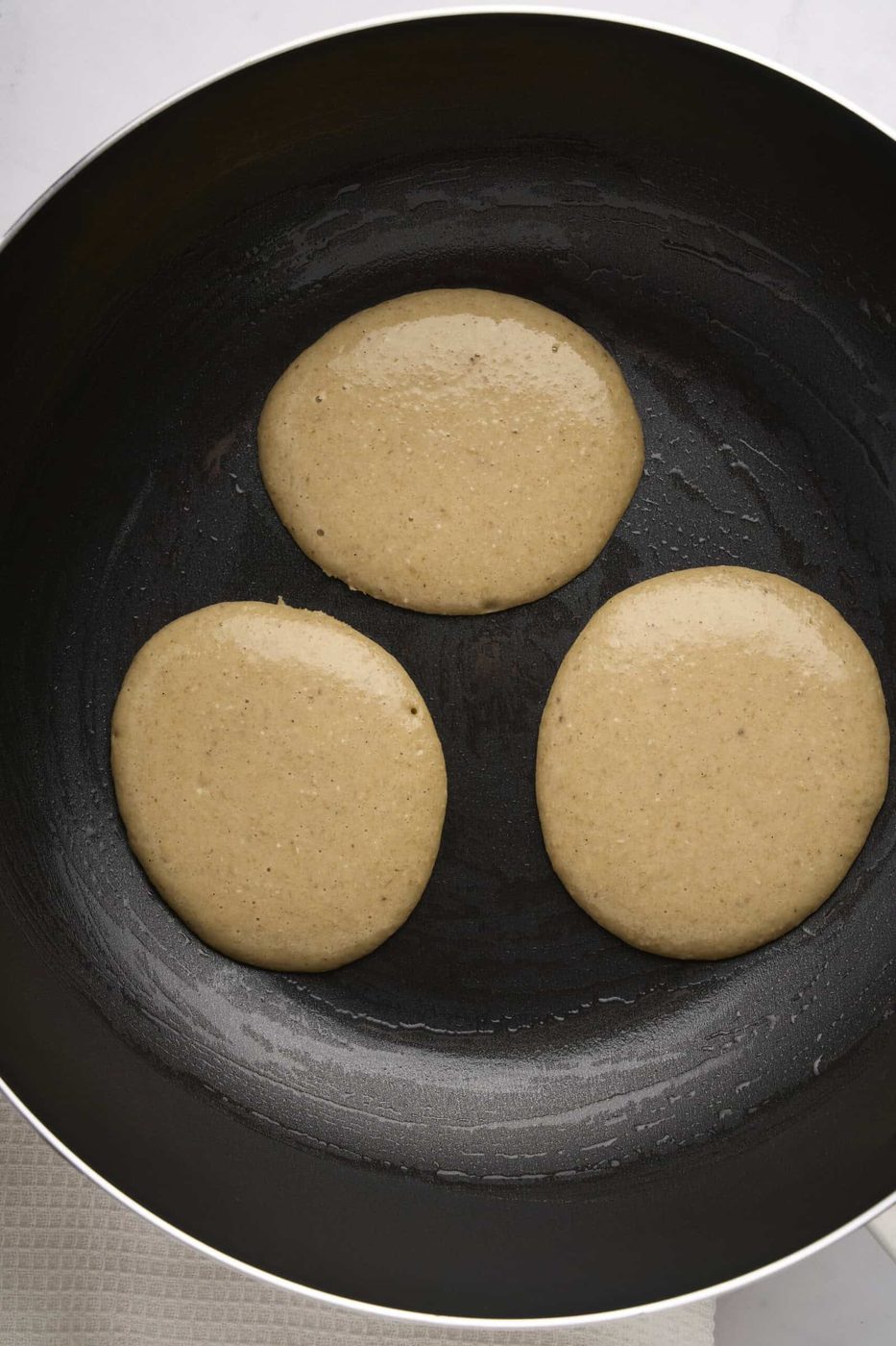 Three round pancakes cooking in a non-stick frying pan on a stovetop. The batter is slightly bubbly, indicating it's being cooked on one side. The pan is black with a smooth surface.