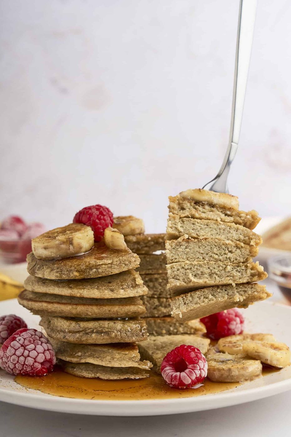 A stack of healthy banana pancakes topped with banana slices, raspberries, and syrup. A fork is lifting a section of the pancakes, showing the fluffy texture. Some frozen raspberries and banana slices are scattered around the plate.