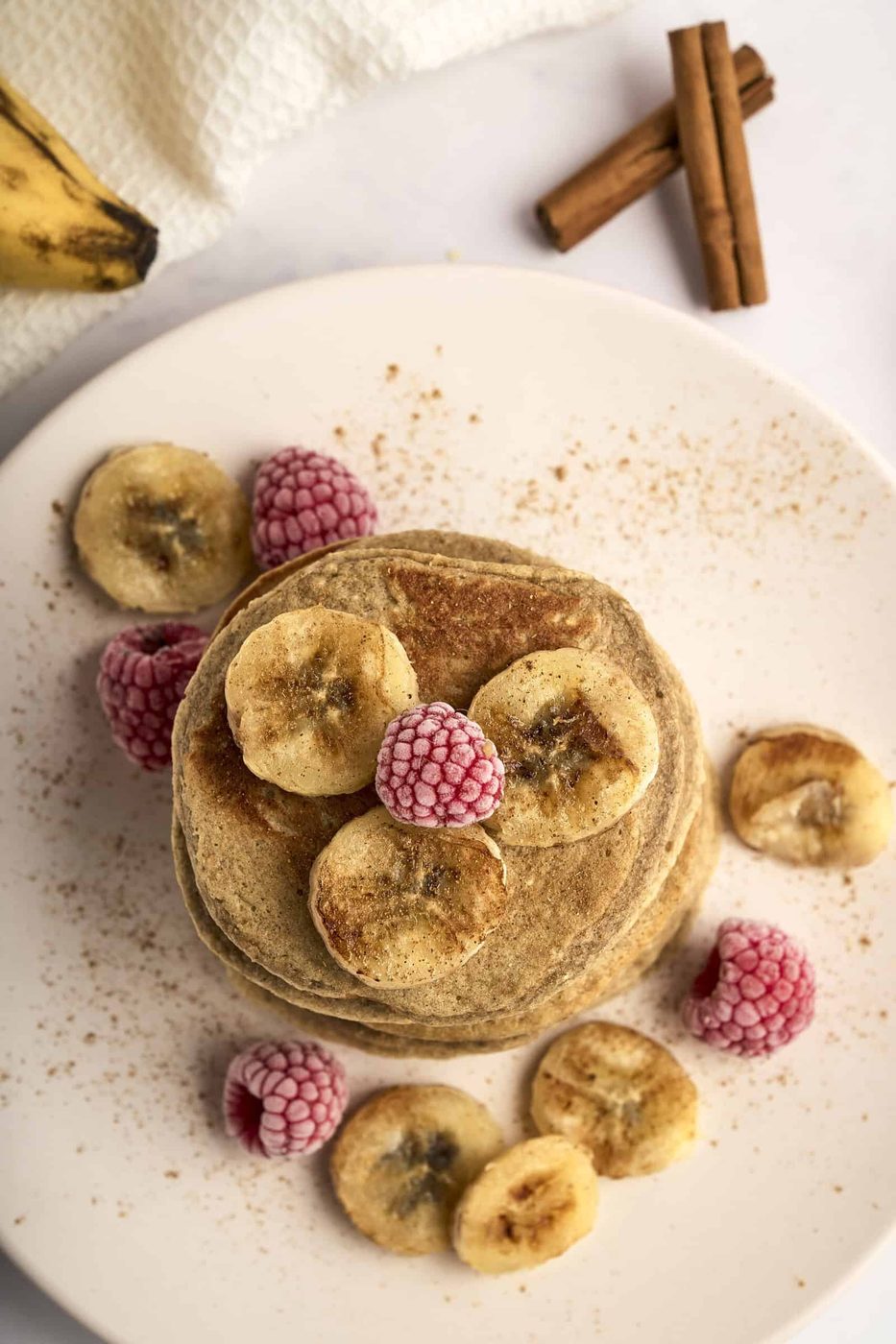 A stack of pancakes topped with banana slices and frozen raspberries on a white plate. Cinnamon sticks are in the background, with a banana and a white cloth nearby. The scene conveys a warm, cozy breakfast setting.