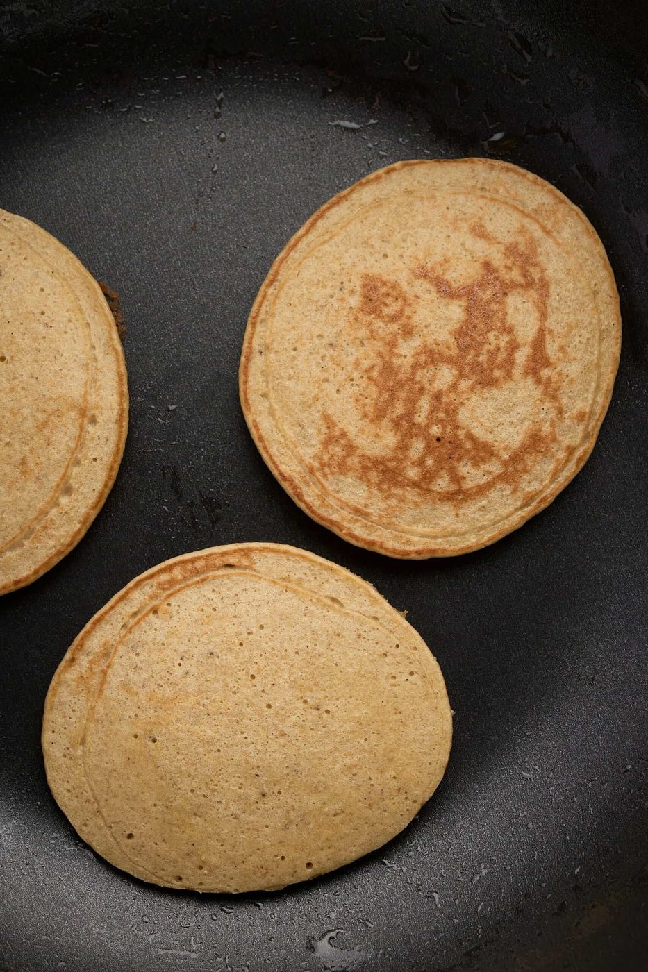 Three golden-brown pancakes cooking in a black skillet. The pancakes have small dark spots and are evenly cooked, indicating they are nearing readiness. The skillet surface is smooth and slightly shiny.