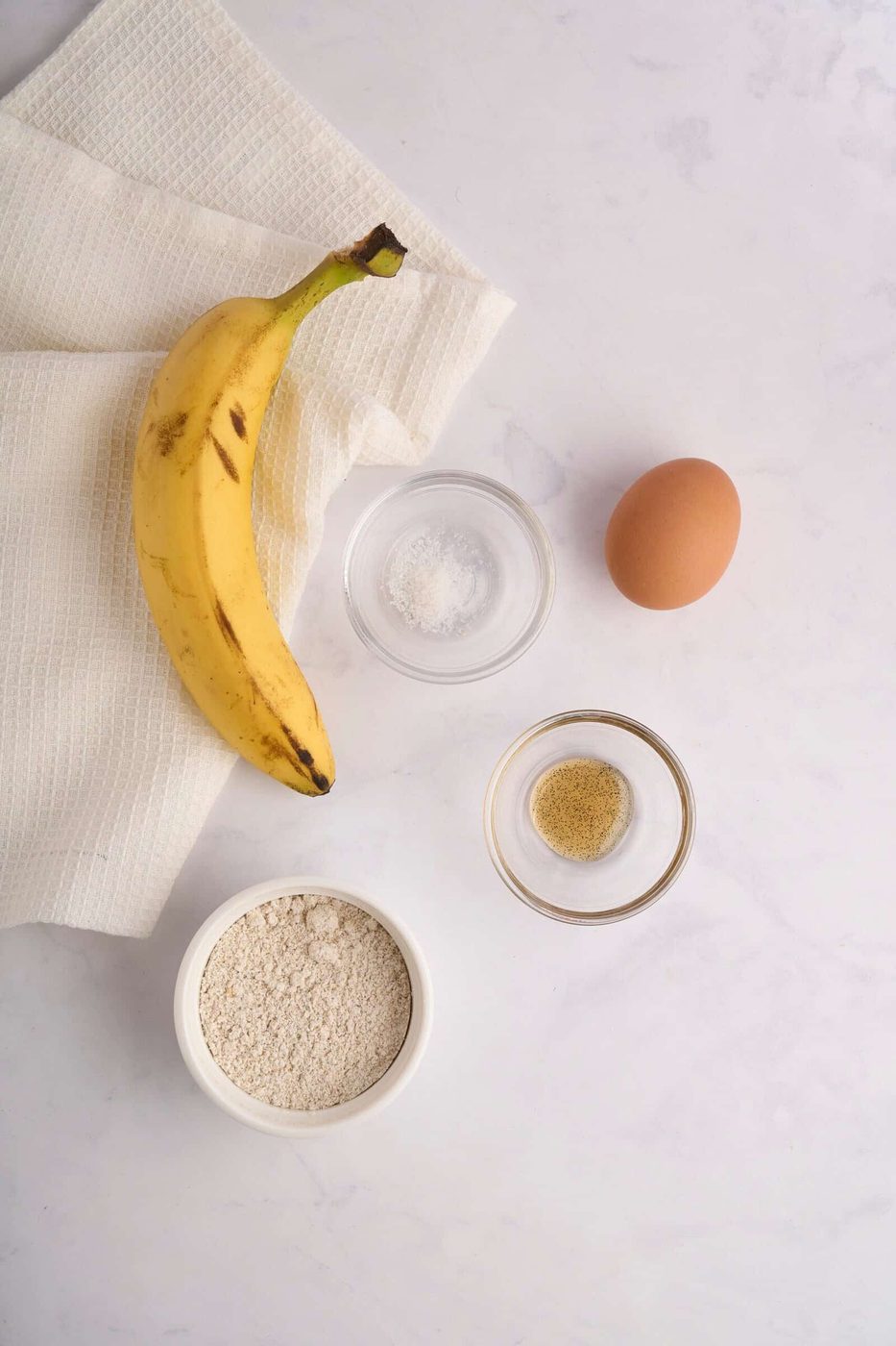 Top view of a banana, an egg, a small bowl of flour, a bowl of white powder (possibly sugar or salt), and a bowl of ground spices on a light surface with a textured cloth. Ingredients suggest preparation for baking or cooking.