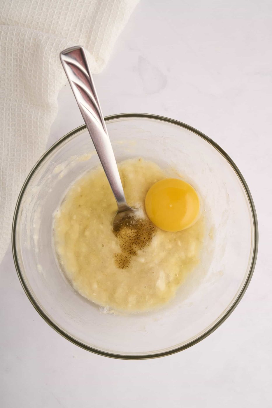 A glass bowl with mashed bananas, a cracked egg, and vanilla extract. A metal spoon rests in the mixture. A white kitchen towel is in the background, set on a marble countertop.