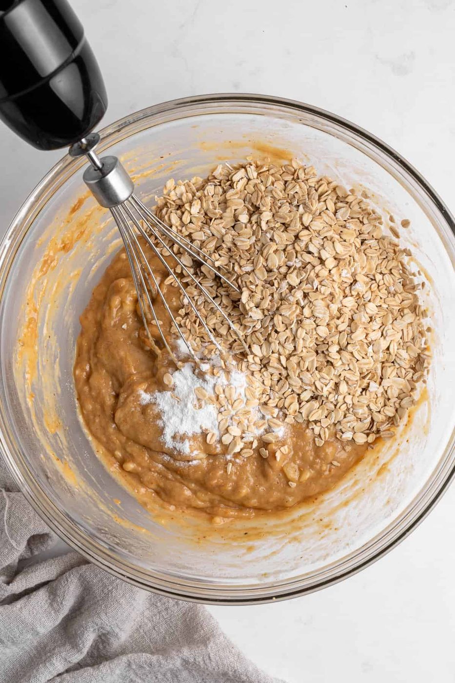 A glass bowl contains a mixture of batter with mashed bananas and oats, being mixed with a whisk. Baking powder is visible on top. The bowl rests on a marble countertop next to a beige cloth.