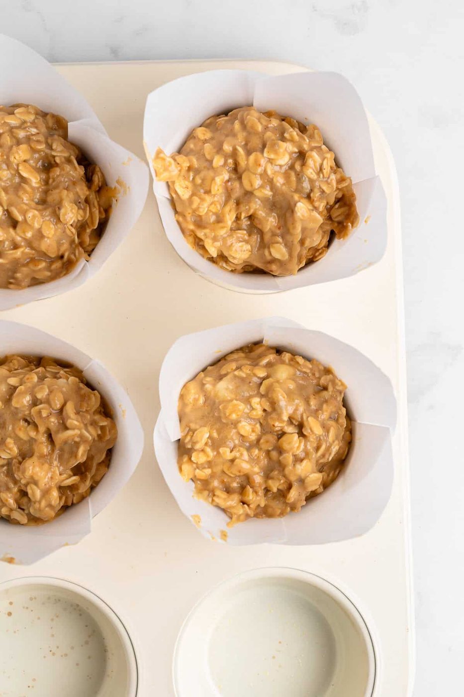 Four Healthy Breakfast Muffins filled with a thick, chunky peanut butter oat batter, set in a white muffin tray. Two of the cups are filled with batter, while the other two compartments are empty, showing a light gray surface.