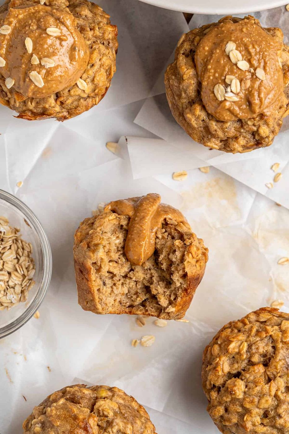 Healthy Breakfast Muffins topped with peanut butter and oats are placed on parchment paper. One muffin is bitten, revealing a moist interior. A small glass bowl with rolled oats is nearby, enhancing the rustic presentation.