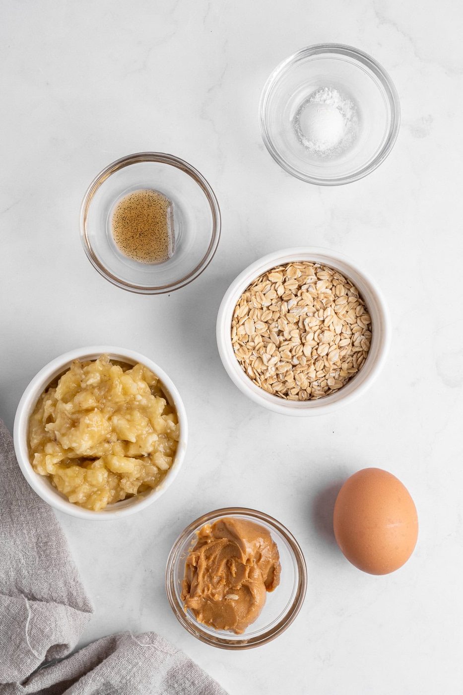Ingredients for a baking recipe are arranged on a white surface. There are bowls containing mashed banana, oats, brown sugar, salt, peanut butter, and a whole egg. A beige cloth is partially visible in the bottom left corner.