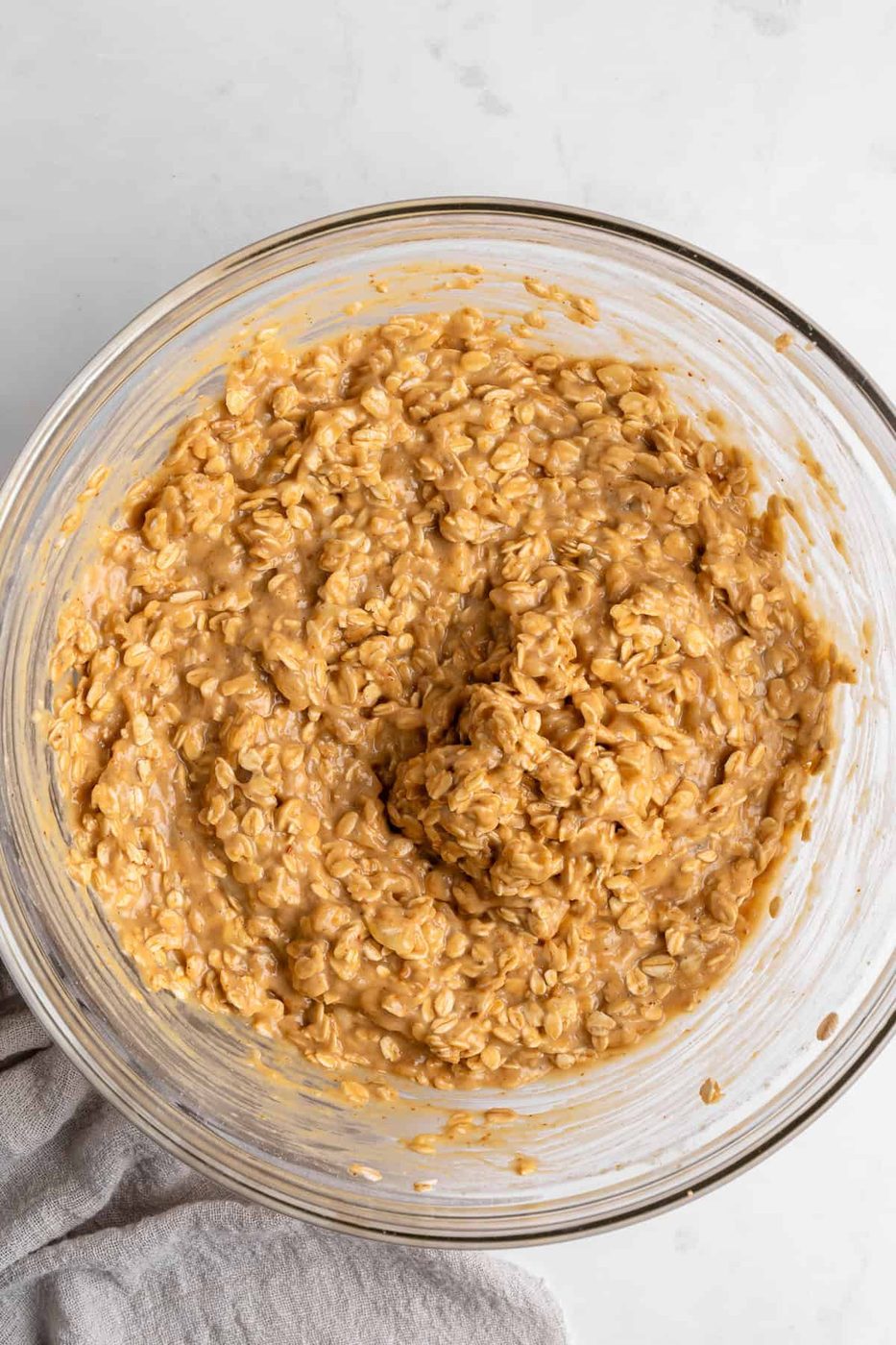 A glass bowl filled with creamy peanut butter oatmeal mixture sits on a white surface. A beige cloth napkin partially appears in the bottom left corner.