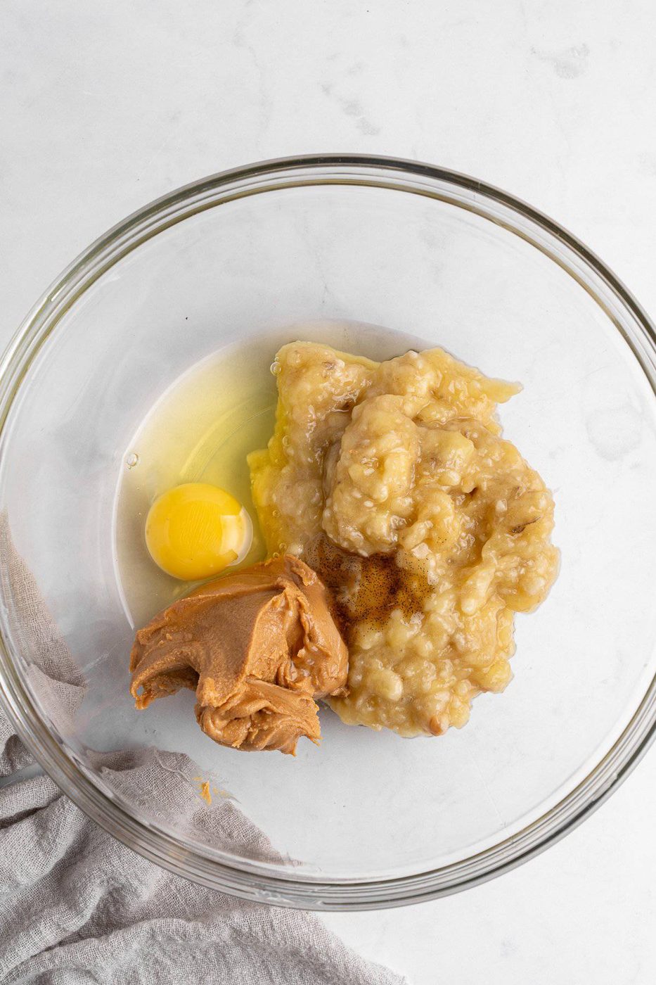 A glass bowl containing mashed banana, a raw egg, peanut butter, and a sprinkle of cinnamon. The ingredients are ready to be mixed, and a light-colored cloth is partially visible on the left side.